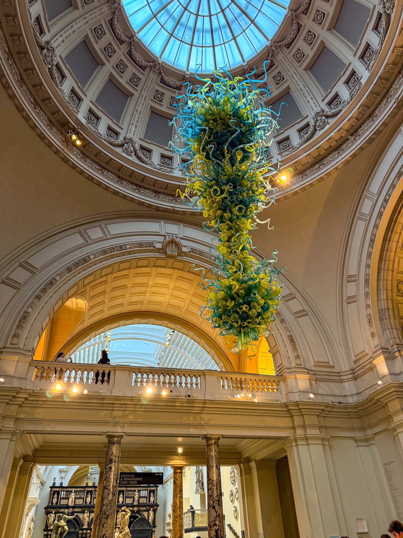 A blue and green glass sculpture hanging in an ornate museum rotunda